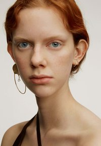 Pendientes de aro dorados con un diseño texturizado y un acento de cristal colgante. El sujeto tiene piel clara y cabello rojo, y lleva un top halter marrón.