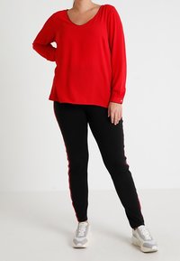 Osoba ubrana w czerwoną bluzę z długim rękawem, czarne spodnie z czerwonymi bocznymi paskami i białe sneakersy, stojąca na tle gładkiej białej ściany.