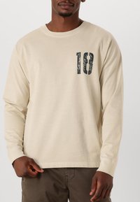 Beiges Langarm-T-Shirt aus Baumwolle mit einem runden Ausschnitt und einer großen, abgenutzten schwarzen "18" auf der linken Brustseite bedruckt.