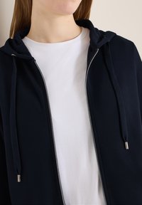 Femme portant un t-shirt blanc à col rond superposé d'un sweat à capuche zippé bleu marine avec cordons de serrage et dents de fermeture éclair en métal.
