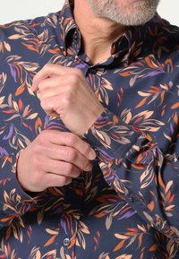 Herenoverhemd met een marineblauwe achtergrond en een bloemenpatroon in oranje, paars en beige. Knopen voor het bevestigen van de manchetten en details van de kraag zijn zichtbaar.