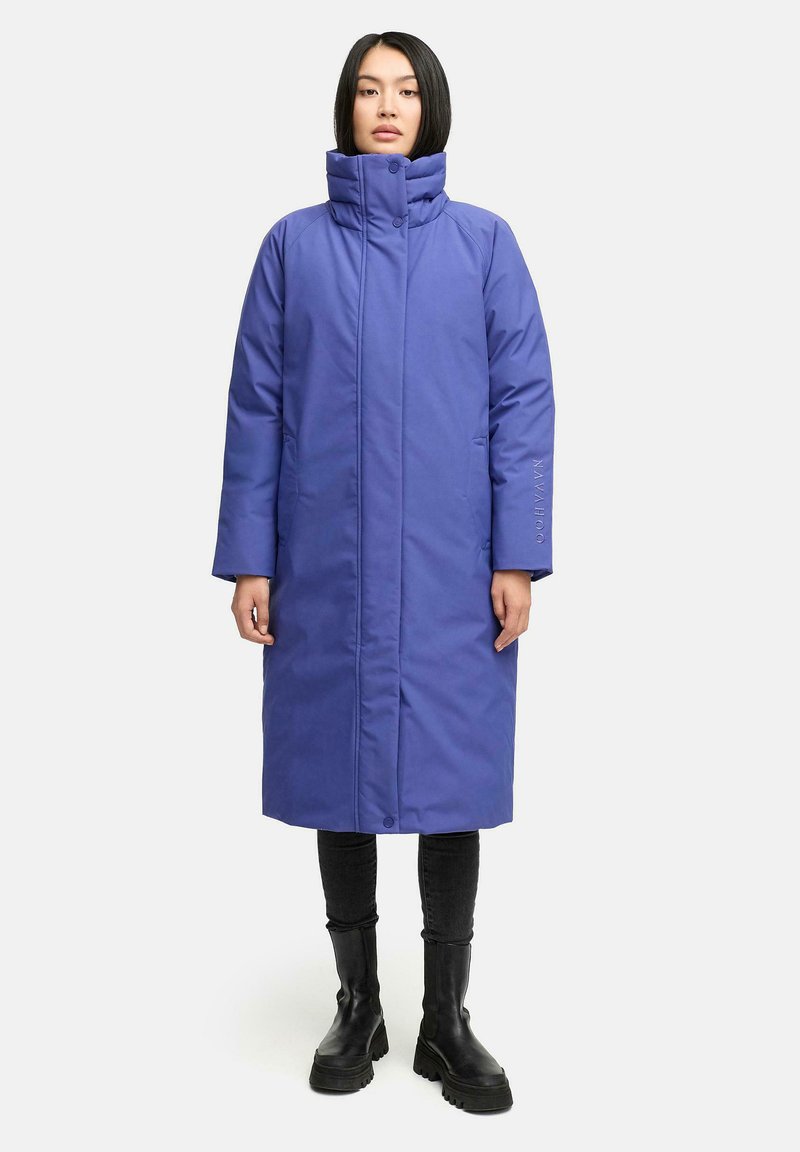 Woman wearing a long, padded, blue winter coat with high collar, black pants, and black chunky boots standing against a white background.