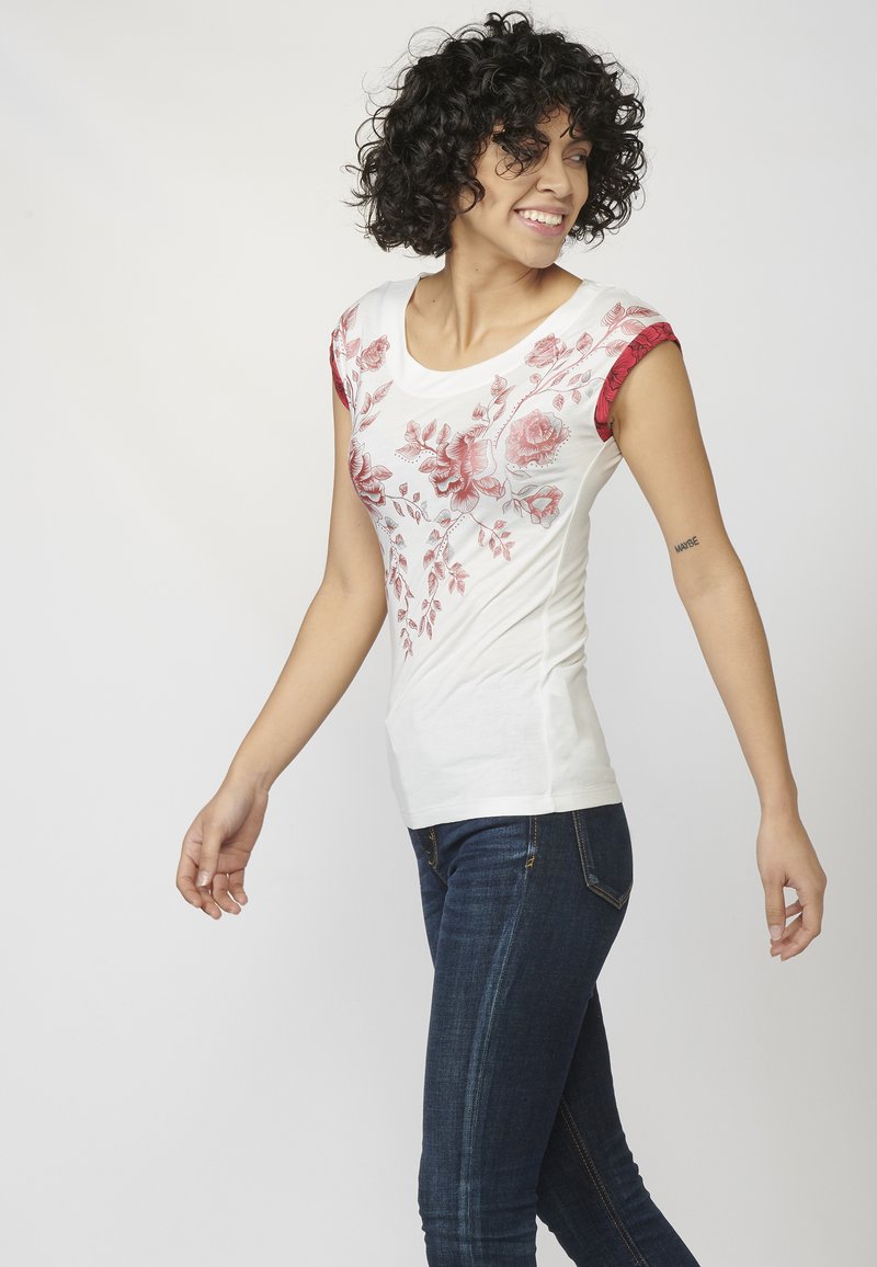 T-shirt blanche ajustée avec broderies florales rouges et manches capuchon rouges à motifs. Assortie avec un jean bleu foncé, mettant en valeur une coupe ajustée.