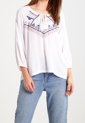 Nainen, jolla on valkoinen löysä paita, jossa on värikästä geometristä kirjontaa ja nauhakuositettu nauha kaula-aukossa, yhdistettynä vaaleansinisiin korkeavyötäröisiin farkkuihin.