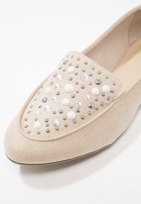 Chaussure plate en daim beige avec un bout arrondi, décorée de clous argentés, de strass transparents et de perles blanches semblables à des perles sur le dessus avant.