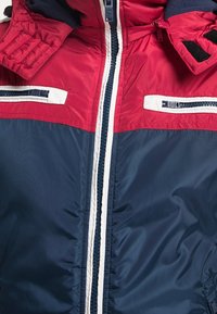 Veste matelassée rouge et marine avec un col haut, une fermeture éclair à l'avant et des accents blancs. Elle dispose de deux poches poitrine zippées. Texture lisse.