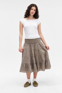 Bílý krátký rukáv top s kulatým výstřihem v kombinaci s hnědou kostkovanou sukní s volány. Modelka nosí olivové slip-on boty a bílé ponožky.