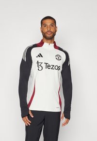 White athletic top with black sleeves, quarter zip, red accents, and "Tezos" logo on the front; detailed Manchester United crest on chest.