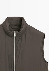 Pikowana bezrękawnik w ciemnoszarym kolorze z wysokim kołnierzem, zamkiem błyskawicznym z przodu i gładką fakturą. Posiada elastyczne wstawki po bokach dla lepszego dopasowania.