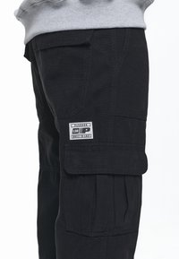 Svarta cargobyxor med flera fickor, synlig sömnad och en etikett med designinslag av "PEGADOR" och "BUILT TO LAST".