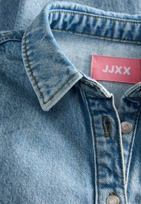 Col de chemise en denim bleu clair avec boutons, surpiqûres marron visibles et une étiquette rose portant l'inscription "JJXX" à l'intérieur de l'encolure.