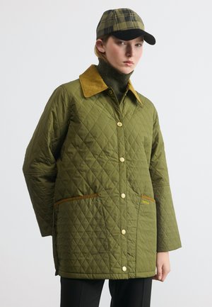 Γυναίκα που φοράει ένα χακί μπουφάν με καπιτονέ επένδυση και καφέ γιακά, μαζί με ένα καρό καπέλο μπέιζμπολ στο ίδιο χρώμα, στέκεται μπροστά σε ένα απλό ανοιχτό φόντο.