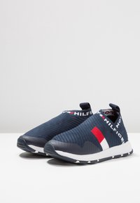 Zapatillas deportivas deslizables de color navy, hechas de un material de punto texturizado. Presentan un logo de acento blanco y rojo en la banda elástica, con una suela blanca y un talón negro.