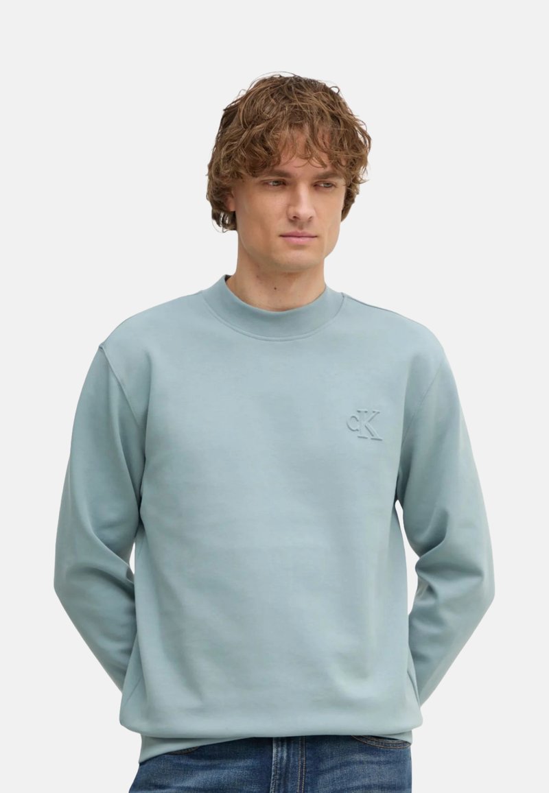 Sudadera azul clara hecha de tela suave, con cuello redondo y logotipo "CK" en relieve en el pecho. Mangas largas y corte relajado.