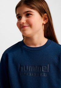 Mörkblå sweatshirt med ribbad halsringning, med en upphöjd "hummel" sportkläder-logotyp med matt finish. Mjukt tyg med en texturerad yta.