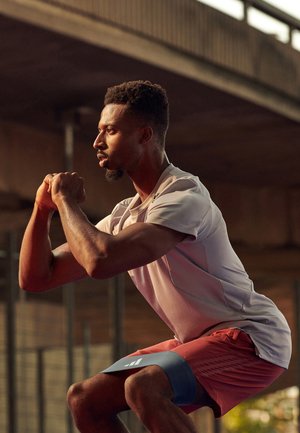Athletic man performing resistance band squats under a bridge, wearing pink shirt and red shorts during outdoor workout.