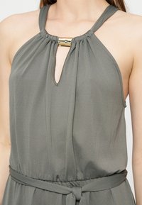 Femme portant une robe grise sans manches à col halter avec un fermoir doré au niveau du décolleté et une ceinture en tissu nouée à la taille.