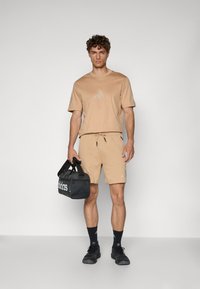 Beige athletic t-shirt and shorts with Adidas logo, paired with black duffel bag and black sneakers. Short sleeves and drawstring waist.