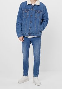 Muž v modré džińové bundě s bílým sherpa límcem, modrých džínách a bílých teniskách, stojí s jednou rukou v kapse.
