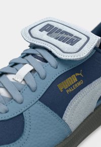 Sinised spordijalatsid, millel on tekstureeritud nahk peal, helesinised ja tumesinised toonid, kullavärviline bränding ning eristuv Velcro rihm.