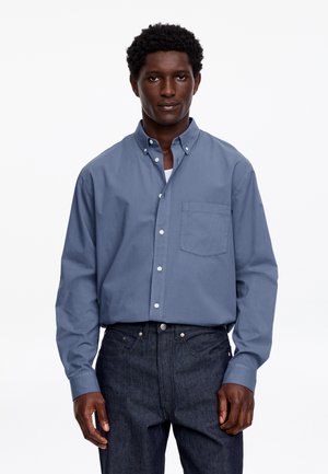 Mężczyzna w niebieskiej koszuli na guziki z długim rękawem wpuszczonej w ciemnoniebieskie dżinsy, stojący na jednolitym białym tle.