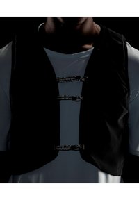 Chaleco negro sin mangas hecho de tela texturizada, que presenta tres broches decorativos plateados en la parte frontal, usado sobre una camisa gris clara.