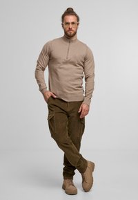 Mies, joka seisoo jalat ristissä, päällä beige vetoketjukauluksellinen neule, oliivinvihreät cargo-housut, vaaleanruskeat nauhakenkäbootit ja kirkasrannekeiset lasit harmaata taustaa vasten.