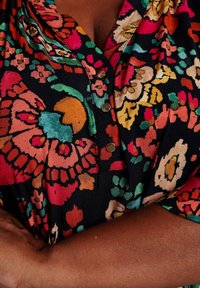 Blusa nera floreale con vivaci motivi in rosa, arancione, verde e crema, dotata di bottoni al colletto e maniche corte. Tessuto morbido.
