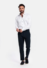 Witte blouse met lange mouwen en donkere knopen, gecombineerd met strakke donkere broek en zwarte instappers, tegen een neutrale achtergrond.