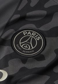 Musta kangas, jossa on maastokuvio. Keskellä oleva pyöreä tägi esittää "PARIS"-logoa, ja "SAINT-GERMAIN"-teksti on alla hopeakirjailulla.