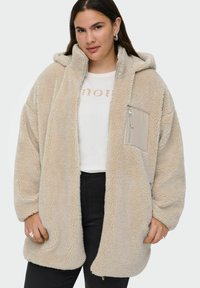 Bežs pārspīlēts fleeces jakas ar kapuci un ar rāvējslēdzēju kabatu, ar mīkstu tekstūru un brīvu piegriezumu. Valkāta virs baltas grafiskās T-kreklu.