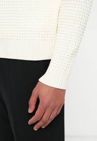 Licht crèmekleurige trui met een getextureerd, verhoogd patroon; geribbelde manchetten en zoom; gedragen met een zwarte broek. Close-up van een hand die op de broek rust.