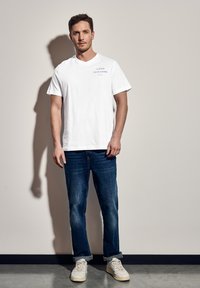 Valkoinen puuvillapaita, jossa on V-pääntie, ja vasemmassa yläkulmassa sinistä tekstiä. Yhdistetty sinisiin farkkuihin ja valkoisiin lenkkareihin. Sileä rakenne.