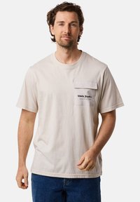 Beige bomullströja med rund halsringning och korta ärmar. Har en framficka med tryckt logotyp på fliken. Bärs med blå jeans.