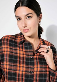 Camicia a quadri nera e arancione in un tessuto morbido, con colletto e bottoni neri. Il modello indossa anelli argentati e una coda di cavallo laterale.