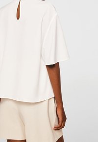 Weißes T-Shirt mit kurzen Ärmeln und einem Schlüsselloch-Ausschnitt am Rücken, aus einem glatten Stoff gefertigt, kombiniert mit hellbeigen Shorts.