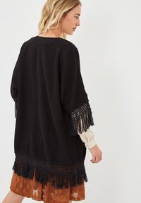 Cárdigan negro de punto con acentos de flecos en las mangas y el dobladillo. Tejido texturizado y corte holgado, superpuesto sobre una blusa de color claro y una falda estampada.