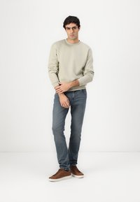 Felpa grigia chiara con polsini e fondo a coste, abbinata a jeans blu e sneakers marroni. Design semplice, texture liscia e colori neutri.