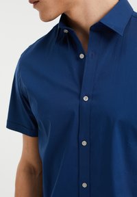 Muž v tmavě modré košili s krátkým rukávem a knoflíky, s límcem, zobrazený od krku po prostředek trupu na bílém pozadí.