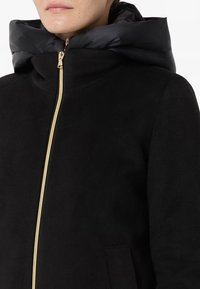Giacca nera con cappuccio imbottito, zip dorata e tessuto testurizzato. Include tasche laterali e un design con colletto alto.