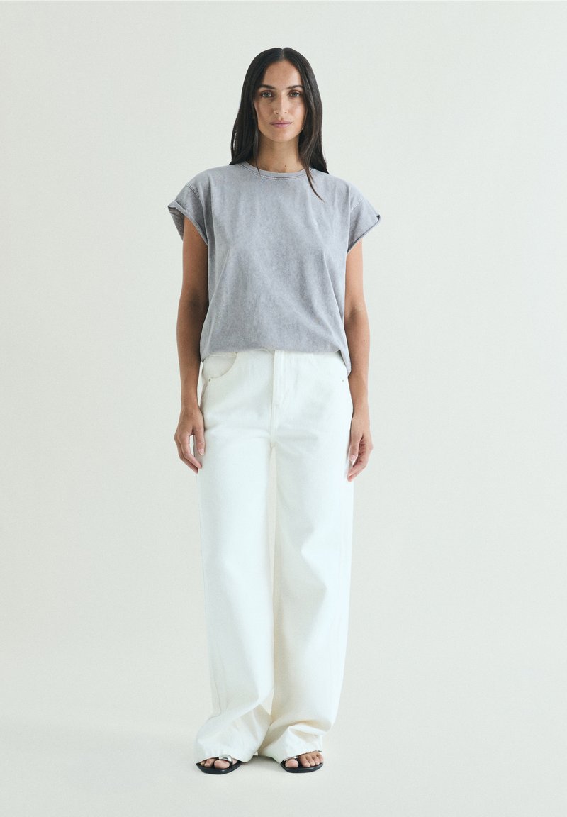 Woman standing wearing a light gray sleeveless top, wide white pants, and black open-toe sandals against a plain light background.
