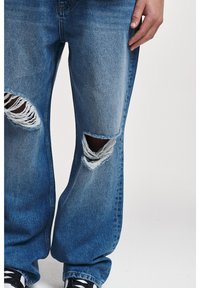 Blue ripped jeans with wide legs, featuring large horizontal tears on both knees, worn with black and white sneakers and a visible hand.