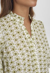 Blusa bianca con ricami floreali verdi, colletto alla cinese e patta dei bottoni. Presenta un tessuto testurizzato e accentuazioni arricciate al colletto.