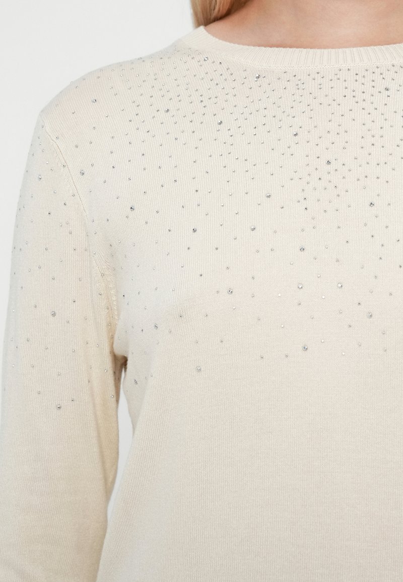 Suéter de punto beige con cuello redondo, tejido texturizado y embellishments plateados dispersos en la sección superior.