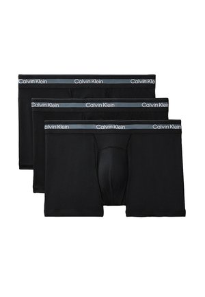 Uomo muscoloso in piedi a piedi nudi su sfondo grigio, indossa boxer neri Calvin Klein, braccia dietro la schiena, volto rivolto in avanti.