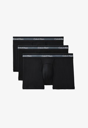 Muskuløs mand stående barfodet mod grå baggrund, iført sorte Calvin Klein boksershorts, med armene bag ryggen, vendt fremad.