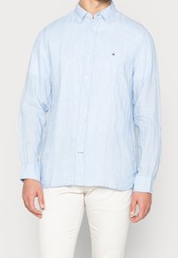 Světle modrá lněná košile s dlouhými rukávy, s límečkem na button-down, předními knoflíky a malým logem na hrudi. Hladká textura.