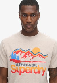 Beige bomulds t-shirt med farverigt bjerg- og solgrafik, med blå træer og rød-orange "Superdry" tekst. Glat tekstur.