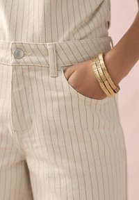 Pantalones a rayas crema con finas líneas rojas; cuenta con un bolsillo lateral, cierre de botón y cremallera. Brazaletes texturados en dorado en la muñeca.