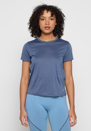 Kobieta z kręconymi włosami, ubrana w dopasowaną niebieską sportową koszulkę z krótkim rękawem i jasnoniebieskie legginsy, stojąca na jednolitym jasnym tle.